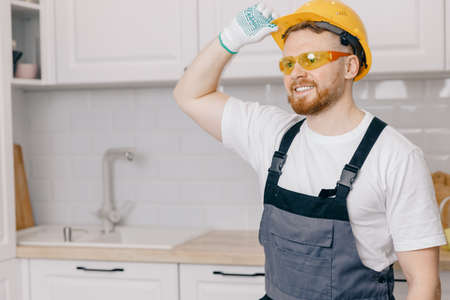 Smile man carpenter worker in protective hard hat stands against background white kitchen. Concept repair and finishing work industry homeの写真素材
