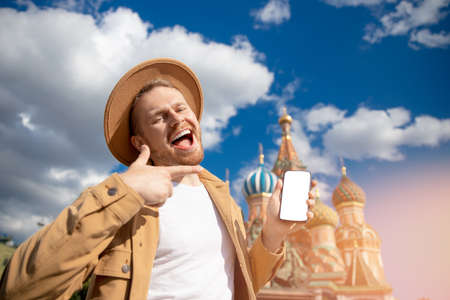 Smile young man tourist in brown hat hold mobile phone background on Kremlin red square in Moscow. Concept travel to Russiaの写真素材