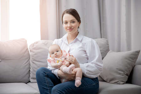 Portrait young mother with newborn baby girl, holding in her arms and sitting in living room on couchの写真素材