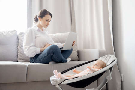 Concept freelance life style maternity leave. Beautiful young mother with baby girl working at home office using laptop computer, sun lightの写真素材