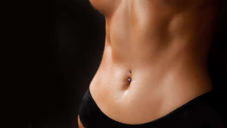 Athletic tanned body of young woman close-up press on black background. Concept of sports, fitness, piercingの写真素材