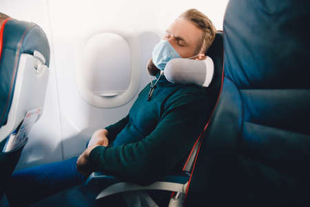 Male passenger in protective mask against viruses of airplane enjoys pillow for sleeping in chair. Concept travel relax planeの写真素材