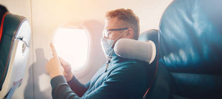 Banner Hipster man in protection mask is watching video on mobile phone, reading online book traveling on airplaneの写真素材