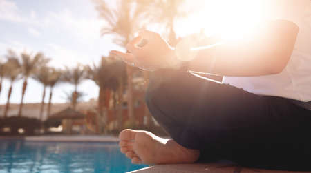 Concept banner asana yoga. Closeup hand of caucasian young man sitting and meditating in lotus pose background blue water and palms sunsetの写真素材