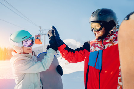 Teamwork winter, friends man and woman high five in alpine skiing equipment with snowboard in their hands against backdrop of snowy mountain and liftsの写真素材