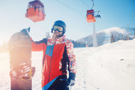 Happy young bearded man with helmet holding snowboard against backdrop of ski resort and lift sunlight. Concept winter sport bannerの写真素材