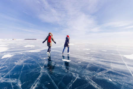 Two young women friends in red cap are skating on ice frozen of Baikal lake, winter sunny dayの写真素材