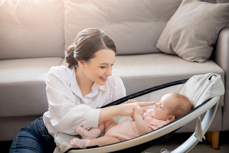 Happy mother playing with girl baby lies in automatic carrycot, sunlight at homeの写真素材
