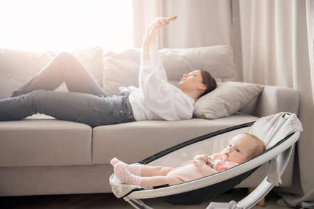 Baby lies in automatic cradle for sleeping, background young mother woman takes selfie on phone. Concept communication and internet work with newbornの写真素材