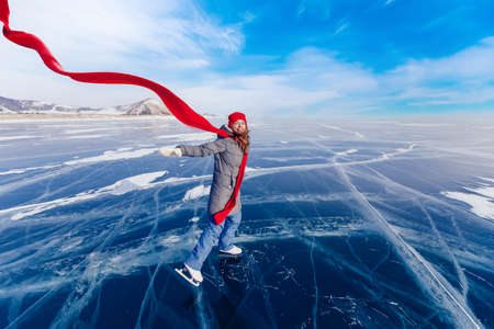 Happy woman tourist on ice skates and with long red scarf on frozen lake Baikal. Concept winter tourism freedomの写真素材