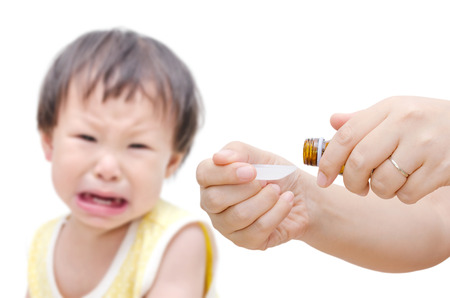 Woman\'s hands pouring medicine in a spoon foreground and crying baby sitting in backgroundの写真素材