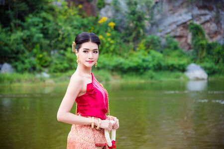 Beautiful asian woman wearing Thai traditional dress holding Thai style flower standing near by the river and smilesの写真素材