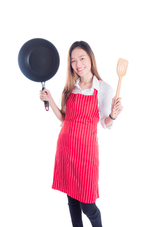 Beautiful young asian housewife wearing red apron , holding pan and turner isolated over white backgroundの写真素材