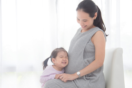 Little asian girl hugging her pregnant mother belly and smiles at homeの写真素材
