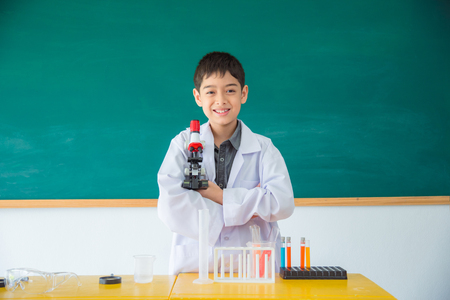 Litlle asian boy wearing white coat and holding microscope smiling in front of classroomの写真素材