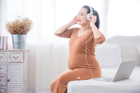 Beautiful asian pregnant woman sitting on bed and listening music from notebook computer via...
