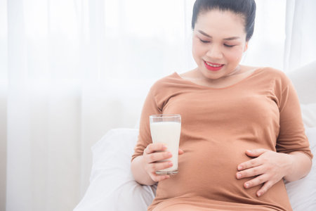 Pregnant woman sitting on bed ,holding a glass of milk and smiles.Focus at milk glass and pregnant belly.の写真素材