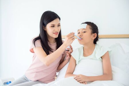 Young asian girl taking syrup medicine from her mother while sitting on bed in bedroomの写真素材