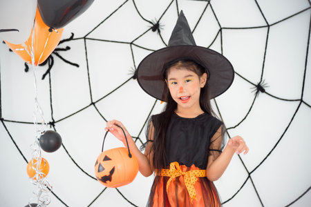 Pretty asian girl in witch costume standing and holding pumpkin bucket in room with Halloween decorationの写真素材