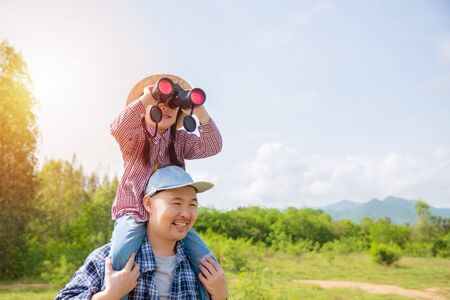 Little asian girl sitting on her father shoulder and looking via binoculars between trekking in forrest.の写真素材