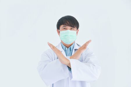 Asian male doctor wearing medical mask showing stop sign with crossed arm over white background.の写真素材