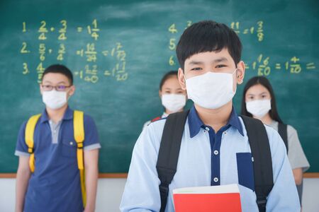 Young asian schoolboy wearing facial mask standing in front of chalkboard and looking at camera,classmate standing on background.の写真素材