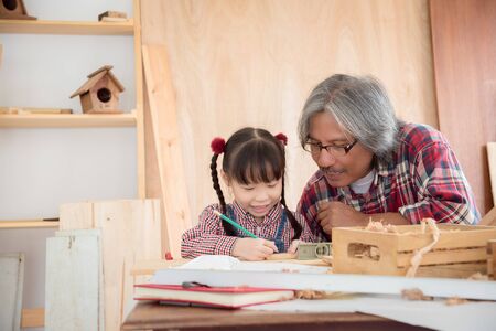 Senior male asian carpenter and grandchild making wooden bird house at home . Little asian girl working at carpentry workshop.の写真素材