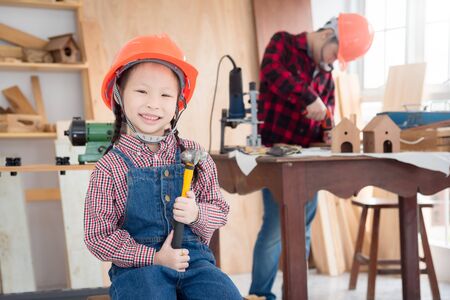 Little asian girl wearing helmet and holding hammer with smiles sitting at carpentry shop.の写真素材