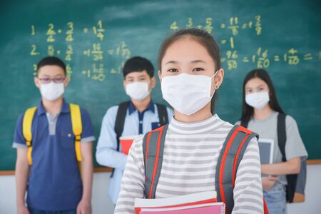 Young asian schoolgirl wearing facial mask standing in front of chalkboard and looking at camera,classmate standing on background.の写真素材