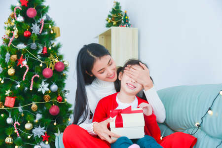 Young Asian mom covering her daughterâs eyes making surprise with a gift box. Near the Christmas tree at home.の写真素材