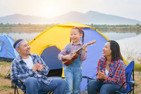 Happy little asian girl playing ukulele and her parents clapping hands at camping site.の写真素材