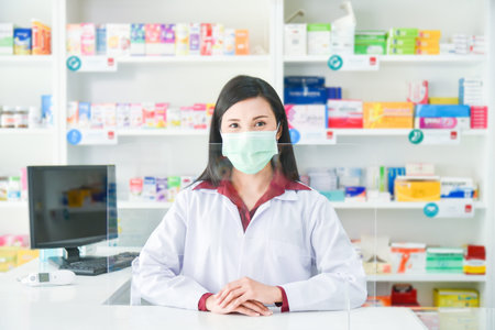 Asian professional young pharmacist wearing face mask while standing behind partition at drugstore.の写真素材