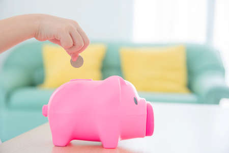 Child hand putting coin to pink piggy bank with sofa background.の写真素材