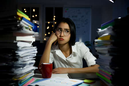 Stressed asian woman sitting at office desk and working overtime with overload work ducument on table.の写真素材
