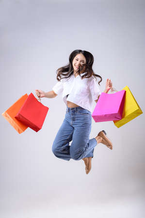 A beautiful Asian woman holding shopping bags of various colors and jumping with a happy expression on a white background. Holiday Shopping.の写真素材