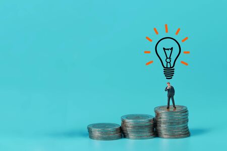 Miniature people: businessman standing on coin stacking podium with light bulb use as backgroundの写真素材