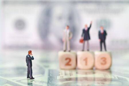 Miniature people: businessman looking and thinking at competitor standing on wooden podium with dollar bank note blur background (Financial and Business competition concept)の写真素材
