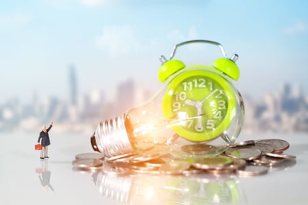 Miniature people: businessman standing beside money and electric glass bulb with city blur background, idea for financial and Business competition conceptの写真素材