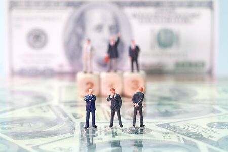 Miniature people: businessman looking and thinking at competitor standing on wooden podium with dollar bank note blur background (Financial and Business competition concept)の写真素材