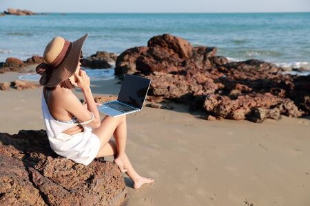 beautiful and cute businesswoman using laptop computer and cell phone with sun glasses and hat on the beach during holiday vacationの写真素材