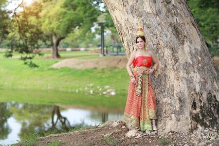 young asian fashion girl wearing Thai traditional costume with beauty face standing beside big green trees in nature outdoors (using for background)の写真素材