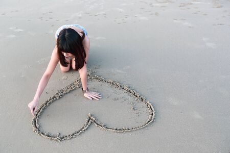 portrait of beautiful woman drawing heart symbol on sandy beachの写真素材