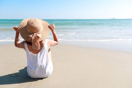 Back view portrait of sexy asian young girl in bikini and hat, looking away to tropical beach while sitting on sandy beach in nature with blue sky.の写真素材