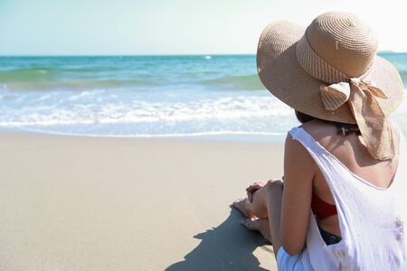 Back view portrait of sexy asian young girl in bikini and hat, looking away to tropical beach while sitting on sandy beach in nature with blue sky.の写真素材