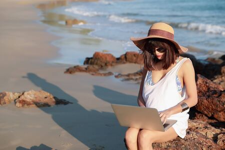 beautiful and cute businesswoman using laptop computer with sun glasses and hat on the beach during holiday vacationの写真素材