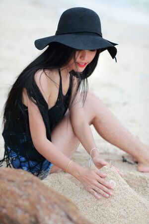 beautiful woman sitting and playing sand on the beach with black hatの写真素材