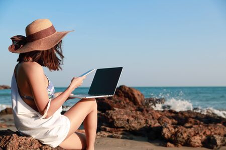 beautiful and cute businesswoman using laptop computer and cell phone with sun glasses and hat on the beach during holiday vacationの写真素材