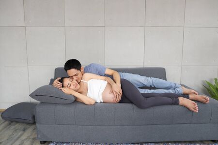 young asian husband lie down on sofa and embracing and kissing pregnant belly wife with love and happiness smile in living roomの写真素材