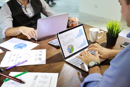 two caucasian businessmen having meeting and using computer with chart on screen and company graph summary report on table with coffee cup and calculator besideの写真素材