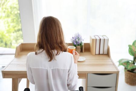 back view of beautiful girl drinking a cup of coffee or tea in the morning with laptop computer and cell phone on wood table in bedroomの写真素材